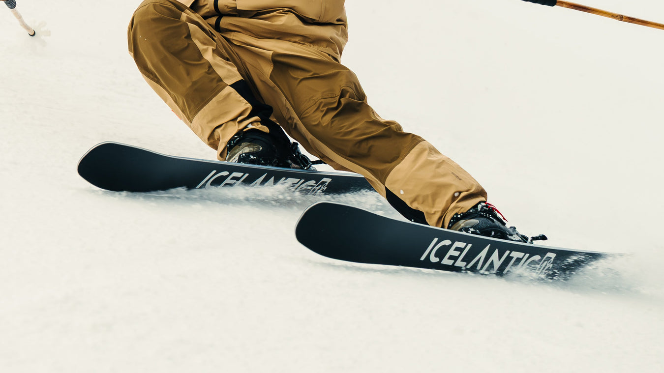 Close-up of a skier in tan pants carving through snow on black skis labeled ICELANTIC, with ski poles visible, creating a spray of snow.