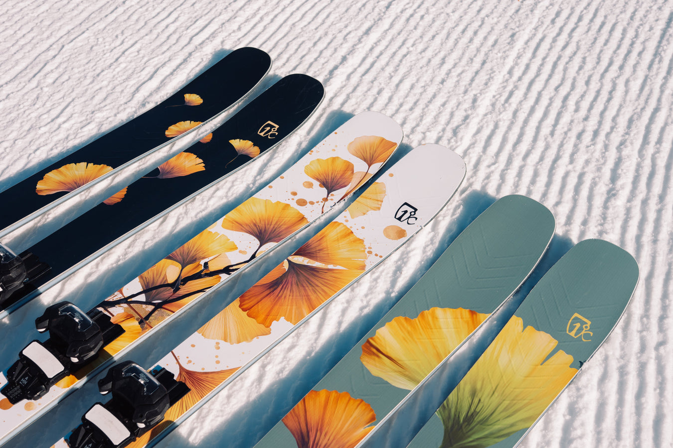 Three pairs of skis with colorful leaf designs lie side by side on groomed snow, featuring shades of black, white, and green with yellow and orange leaf patterns.