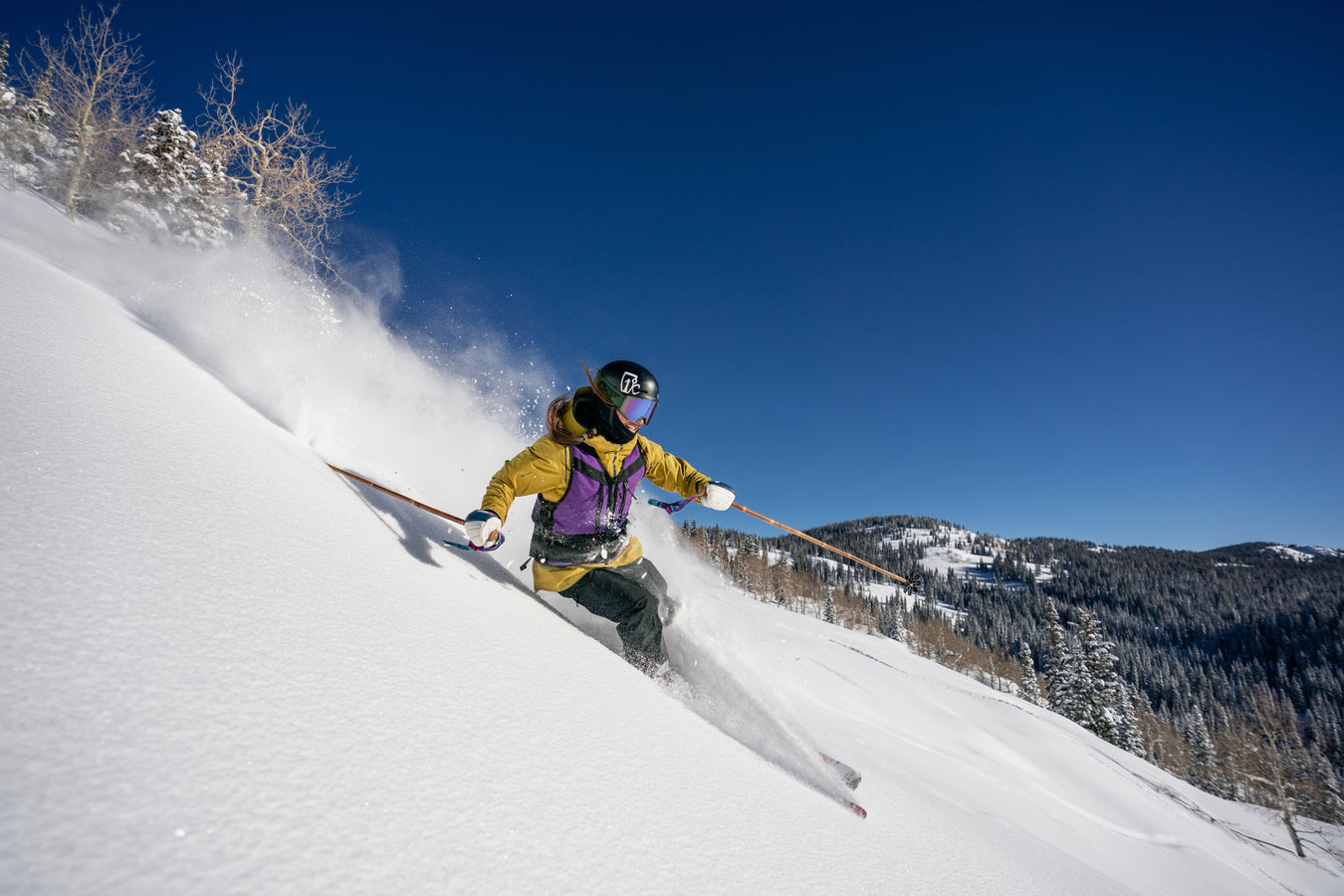 A skier carves through powder snow on a steep slope, while two colorful skis stand upright in deep snow next to snow-covered trees.