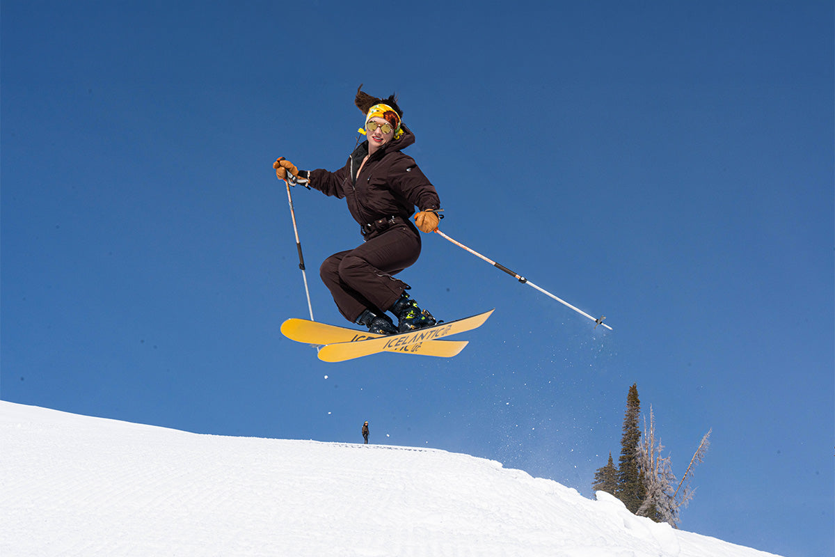 A person in a brown ski suit and yellow earmuffs jumps on yellow skis against a clear blue sky, holding ski poles, with snow and trees in the background.