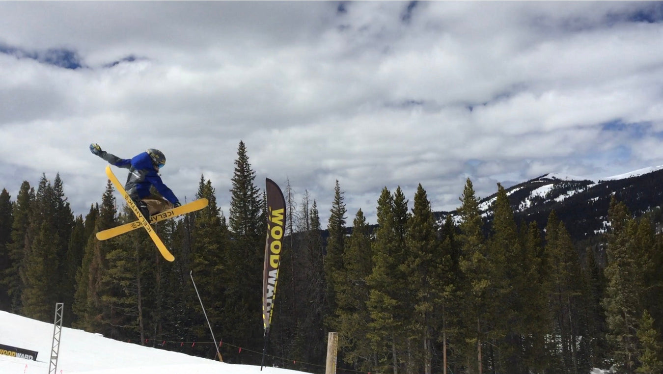 A skier in a blue jacket jumps high in the air, crossing yellow skis, with pine trees, a Woodward banner, and snow-covered mountains in the background under a cloudy sky.