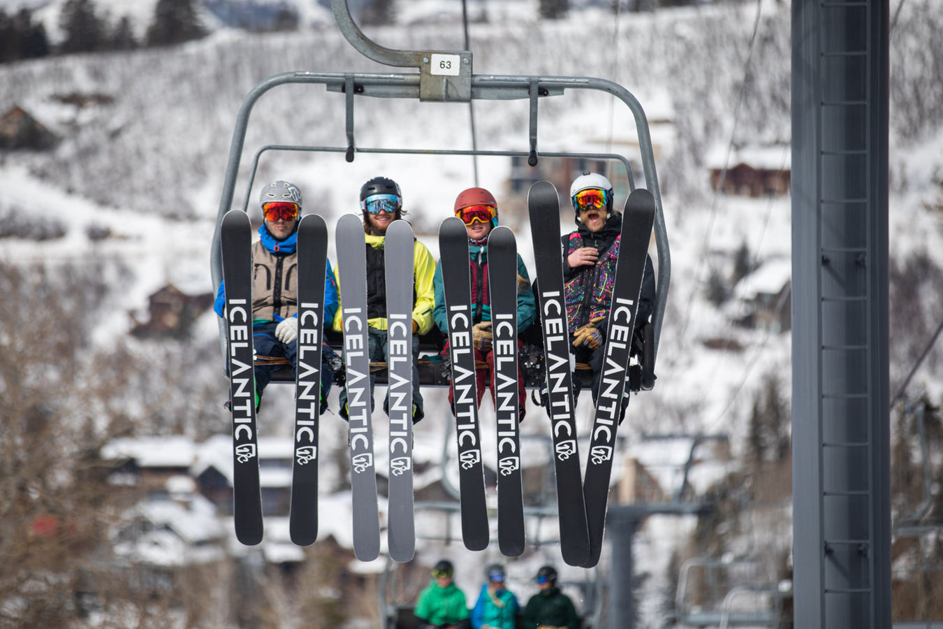 Four skiers in winter gear sit on a ski lift, their skis labeled Icelantic hanging down. Snowy landscape and houses are visible in the background. Two more skiers in green are below.