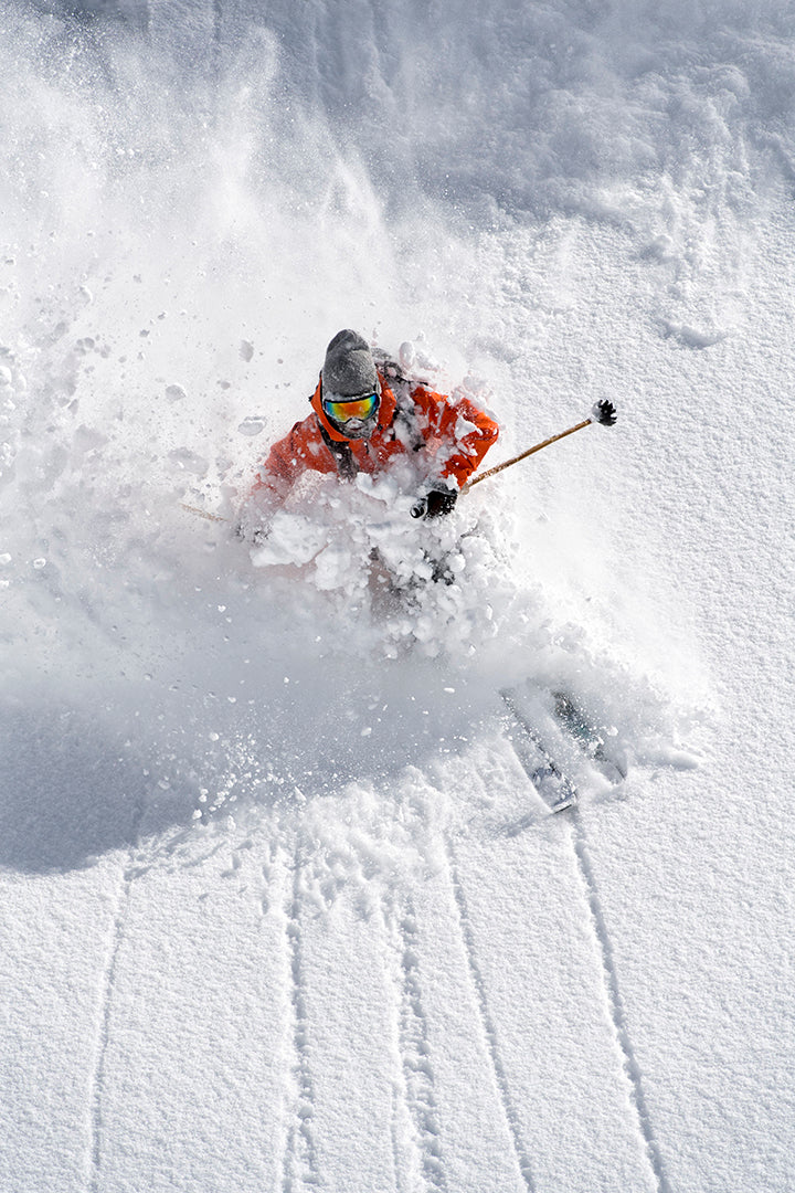 A skier in an orange jacket and gray helmet skis through deep powder snow, creating a spray of snow around them on a bright, sunny day.