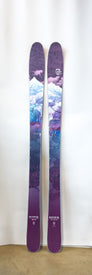 A pair of Demo - 23/24 Riveter 85 women’s skis by Icelantic, featuring artwork of snow-capped mountains, clouds, and a starry sky in purple tones, stand upright against a white background—an all-mountain ski crafted for both style and performance.