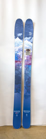 A pair of blue Icelantic Demo - 23/24 Riveter 104 all-mountain women's skis with a mountain and sky design—featuring snowy peaks, clouds, and rocky terrain—are standing upright against a plain white background.