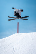 A skier riding the Icelantic Skis Nomad 100 catches air while performing a trick over a red pole on a snowy slope, set against a clear blue sky.