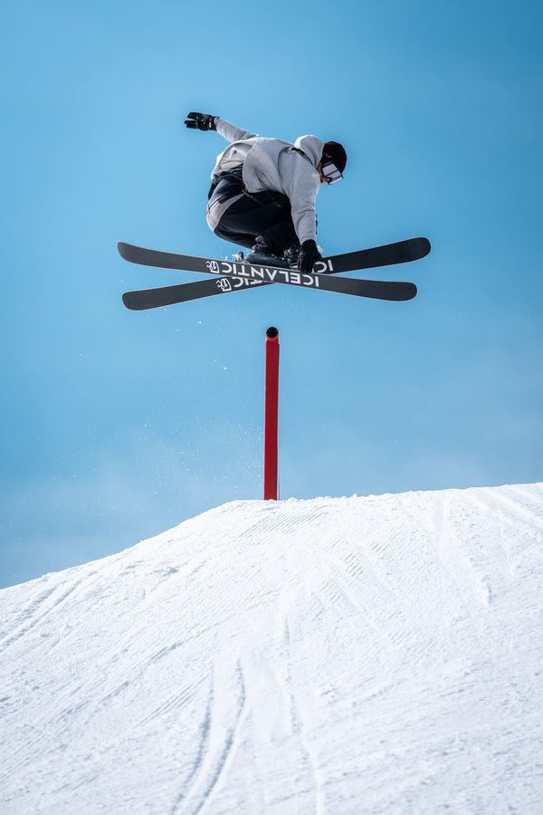 A skier riding the Icelantic Skis Nomad 100 catches air while performing a trick over a red pole on a snowy slope, set against a clear blue sky.