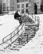 A skier equipped with Icelantic Skis’ Nomad 94 glides down a snow-covered handrail beside an outdoor staircase, as another person waits by the entrance holding a camera.