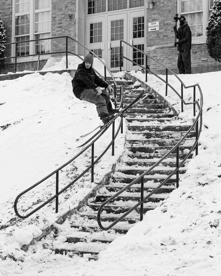 A skier equipped with Icelantic Skis’ Nomad 94 glides down a snow-covered handrail beside an outdoor staircase, as another person waits by the entrance holding a camera.