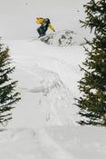 Soaring off a snowy slope on Icelantic Skis’ Nomad 106, a skier in a yellow jacket catches air, framed by snow-covered trees and mountains in the background.