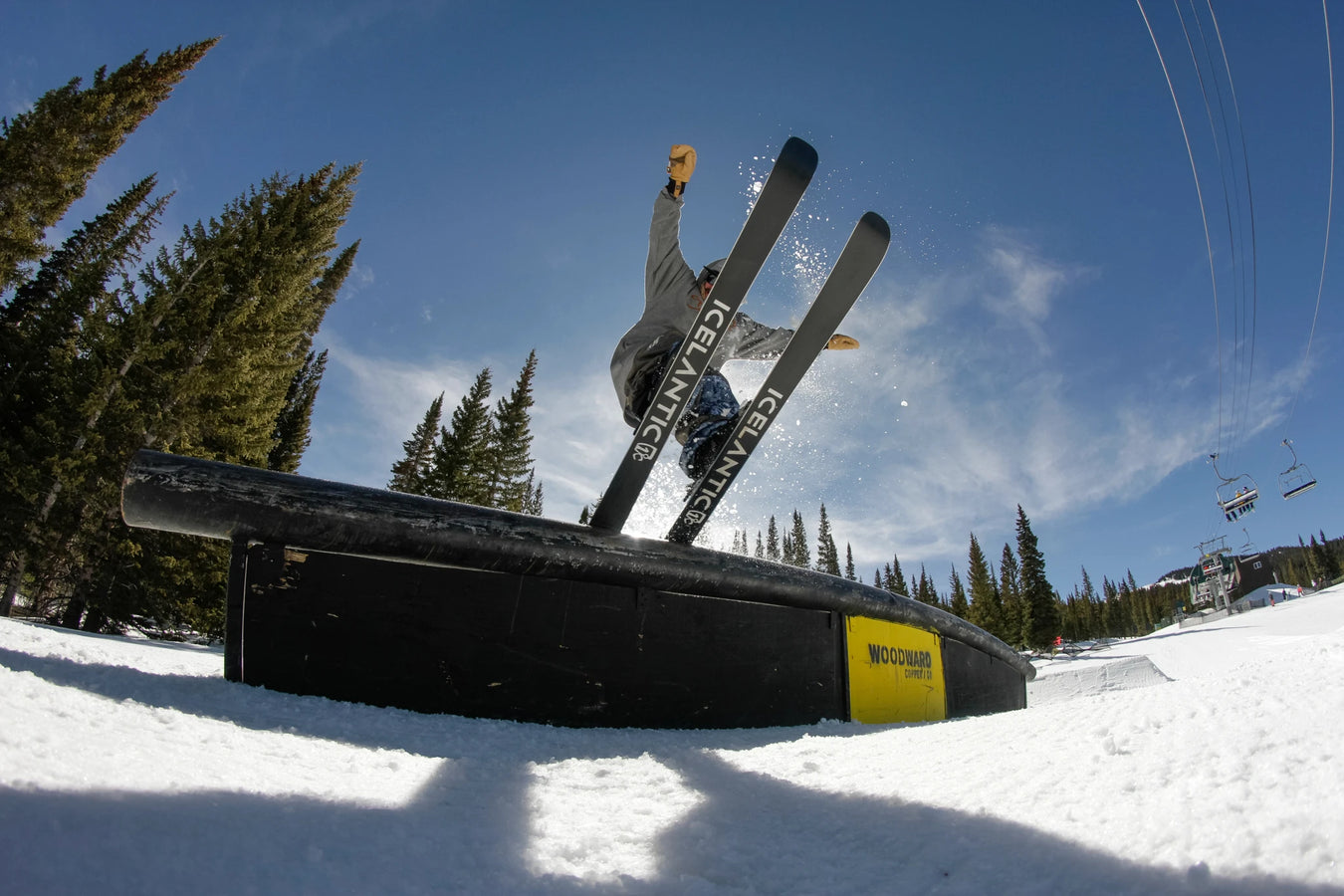 A skier rides the Icelantic Skis Nomad 94 on a rail in a snowy terrain park, with trees and a ski lift in the background beneath a clear blue sky.