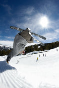 A skier catches air on a snowy slope, performing a flip trick on the Icelantic Skis Nomad 94 under the bright sun, with trees, mountains, and other people visible in the background.