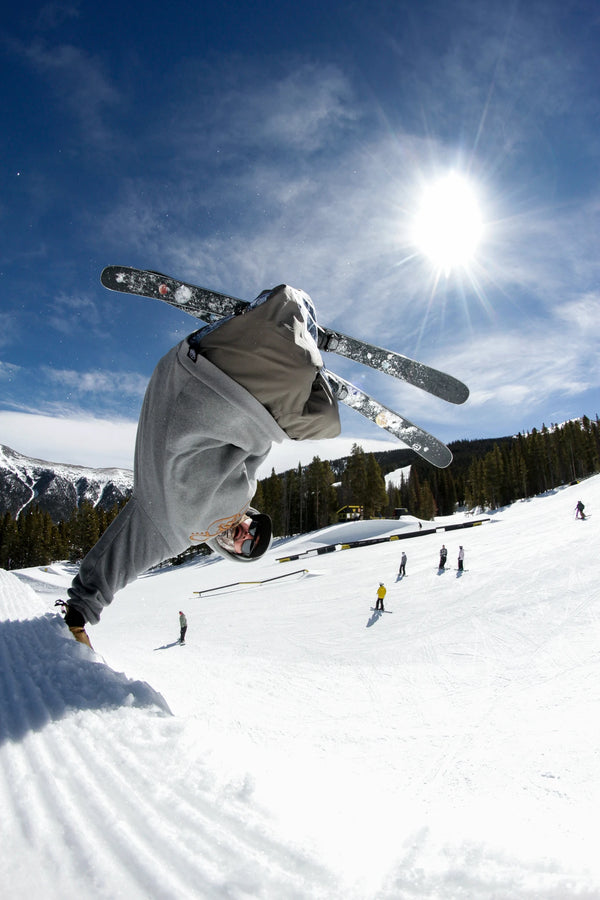 A skier catches air on a snowy slope, performing a flip trick on the Icelantic Skis Nomad 94 under the bright sun, with trees, mountains, and other people visible in the background.
