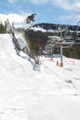 Soaring through the air on a sunny day, a skier performs a trick off a snowy jump near a ski lift, with pine trees and mountains in the background, riding the Icelantic Skis Nomad 100.