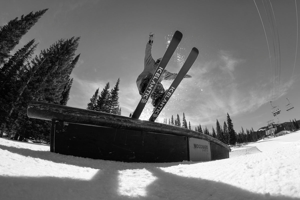 A skier slides on a rail in a snowy terrain park, with trees and a ski lift in the background, captured from a low angle in black and white.