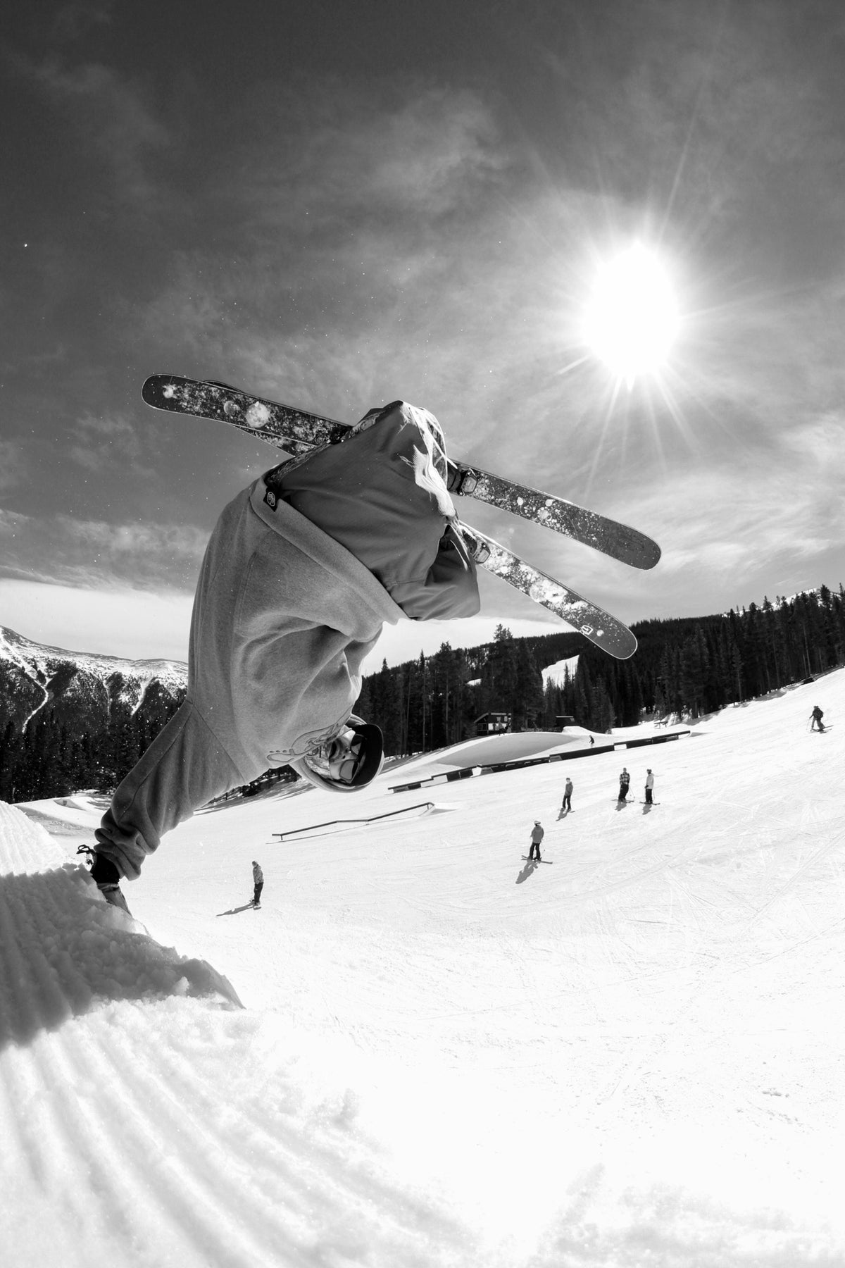 A skier in mid-jump performs a trick on a snowy slope under a bright sun, with other skiers and trees visible in the background.
