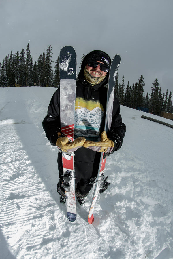 A skier dressed in winter gear smiles while standing on snow, holding two upright Icelantic Skis Nomad 100 skis and a clear trophy; they’re wearing sunglasses and gloves, with pine trees and a cloudy sky in the background.