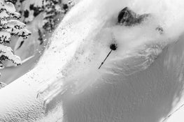A skier speeds downhill through deep powder on a steep, snow-covered mountain slope surrounded by trees, riding the Icelantic Skis Nomad 112 and sending up a plume of fresh snow.