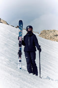 Clad in black gear and a helmet, a skier stands on a snowy slope with rocky terrain in the background, holding an upright pair of Icelantic Skis Maiden 94 in one hand and a ski pole in the other.
