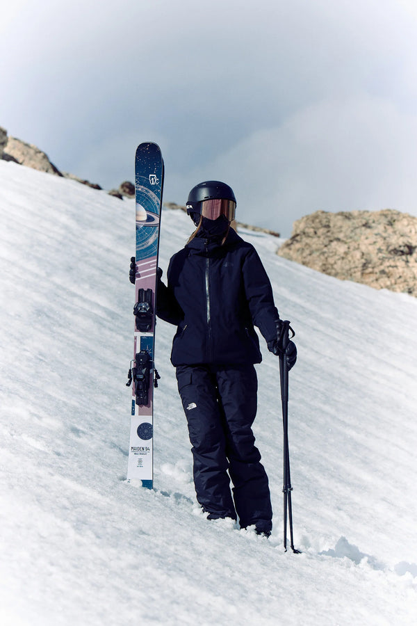 Clad in black gear and a helmet, a skier stands on a snowy slope with rocky terrain in the background, holding an upright pair of Icelantic Skis Maiden 94 in one hand and a ski pole in the other.