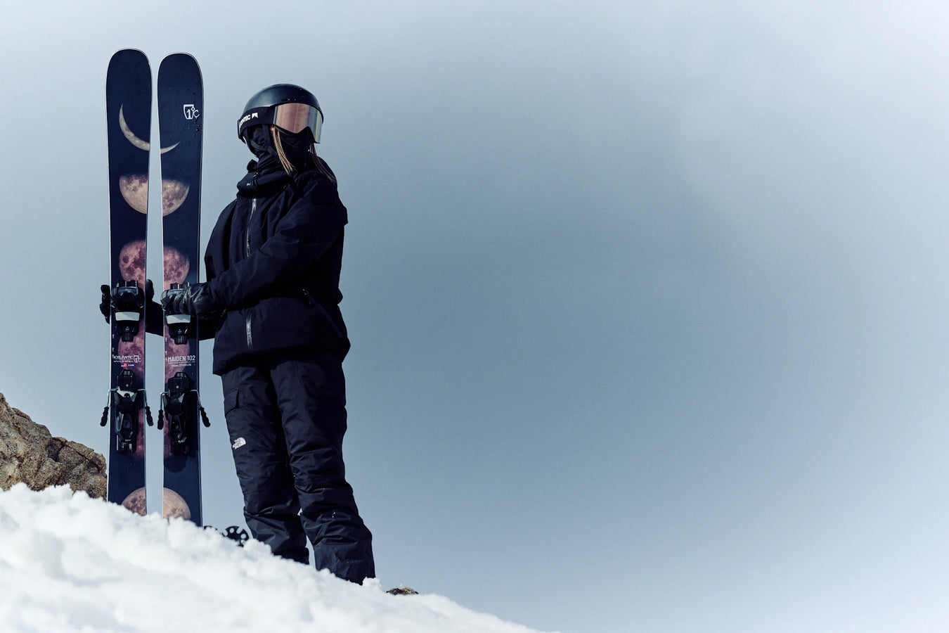 Dressed in black ski gear and a helmet, a person stands on snowy ground against a cloudy sky, holding a pair of Icelantic Skis Maiden 102 featuring moon designs.
