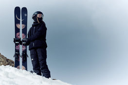 Dressed in black ski gear and a helmet, a person stands on snowy ground against a cloudy sky, holding a pair of Icelantic Skis Maiden 102 featuring moon designs.