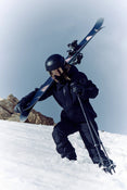 Clad in black gear and helmet, a skier carries Icelantic Skis’ Maiden 102 on their shoulder while climbing up a snowy slope with ski poles under a clear sky.
