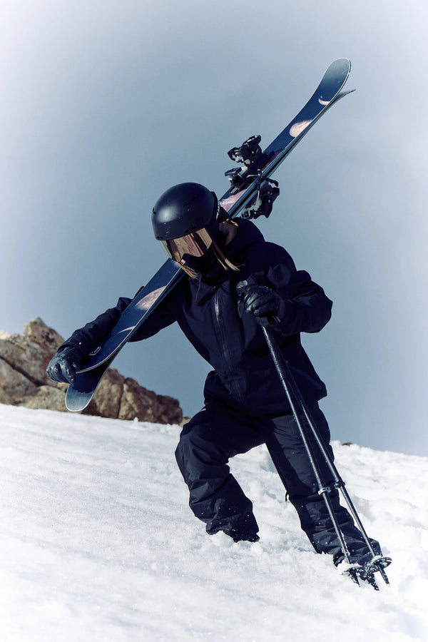 Clad in black gear and helmet, a skier carries Icelantic Skis’ Maiden 102 on their shoulder while climbing up a snowy slope with ski poles under a clear sky.