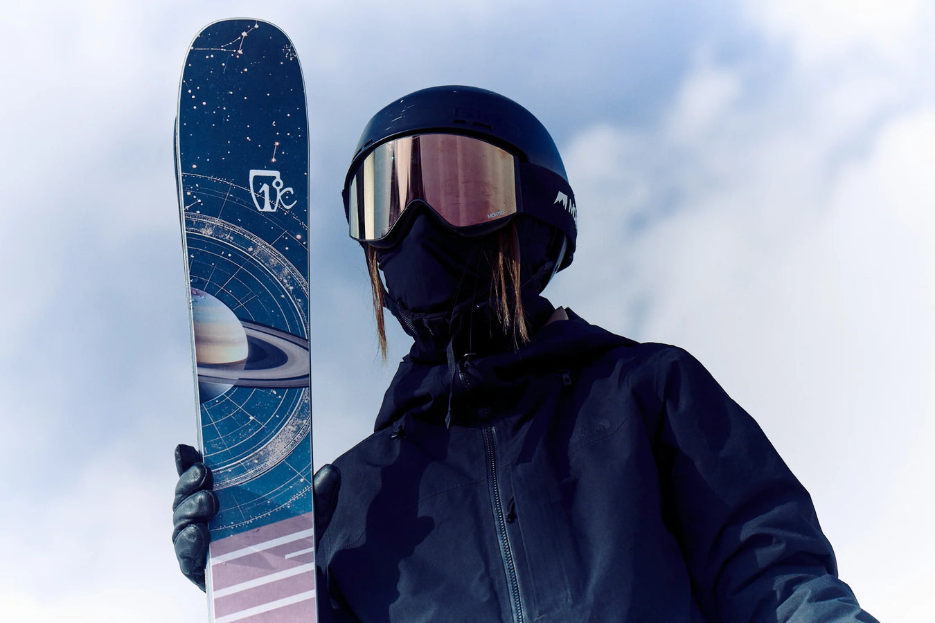 Wearing dark gear and reflective goggles, a skier holds up the Maiden 94 ski by Icelantic Skis, which features a space-themed design with Saturn and orbital lines, all set against a cloudy sky.