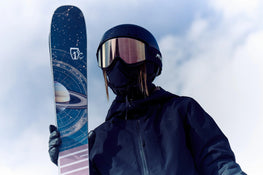 Wearing dark gear and reflective goggles, a skier holds up the Maiden 94 ski by Icelantic Skis, which features a space-themed design with Saturn and orbital lines, all set against a cloudy sky.