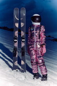 Wearing a shiny pink spacesuit and helmet, a person stands on snowy ground beside a pair of Icelantic Skis’ Maiden 102 skis, which are decorated with moon phases, with a dark sky in the background.