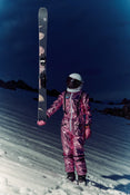 A person in a shiny pink spacesuit and helmet stands on snowy ground at night, holding an Icelantic Skis Maiden 102 vertically with one hand, while dark mountains rise in the background.