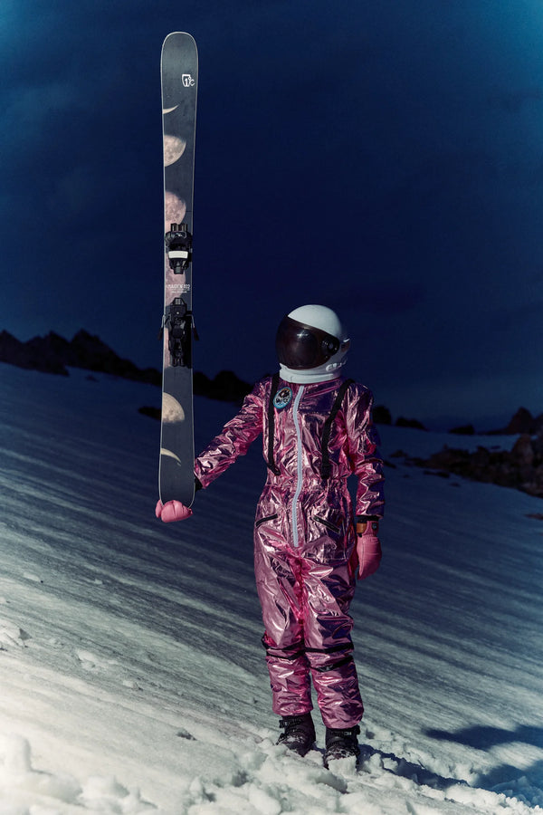 A person in a shiny pink spacesuit and helmet stands on snowy ground at night, holding an Icelantic Skis Maiden 102 vertically with one hand, while dark mountains rise in the background.