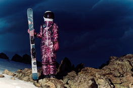 Clad in a metallic pink spacesuit and helmet, a figure stands on rocky ground holding the Icelantic Skis Maiden 94 against a snowy landscape, beneath a dark, cloudy sky.