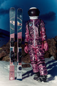 Wearing a metallic pink spacesuit and helmet, a person stands on snow beside the Icelantic Skis Maiden 94, which feature a space-themed design, set against a rocky twilight mountain backdrop.