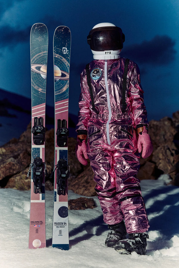 Wearing a metallic pink spacesuit and helmet, a person stands on snow beside the Icelantic Skis Maiden 94, which feature a space-themed design, set against a rocky twilight mountain backdrop.