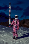 Wearing a shiny pink spacesuit and helmet, a person stands on snow at dusk, holding an upright Icelantic Skis Maiden 94. Rocky terrain and a dark blue sky with clouds create a dramatic backdrop.