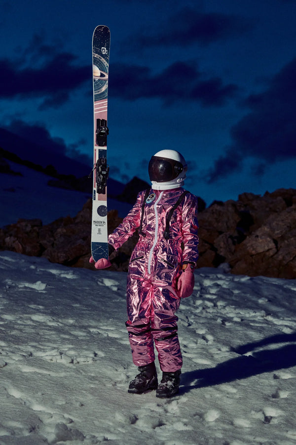 Wearing a shiny pink spacesuit and helmet, a person stands on snow at dusk, holding an upright Icelantic Skis Maiden 94. Rocky terrain and a dark blue sky with clouds create a dramatic backdrop.
