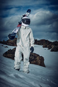 Clad in a white suit and helmet, an individual carries the Icelantic Skis Nomad 100 on their shoulder while standing on snowy terrain dotted with rocky outcrops beneath a cloudy sky.