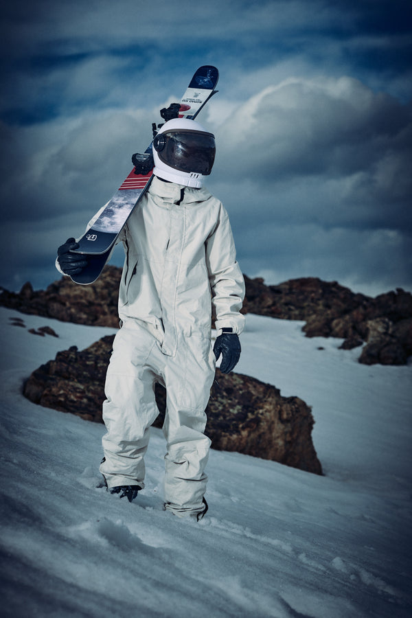 Clad in a white suit and helmet, an individual carries the Icelantic Skis Nomad 100 on their shoulder while standing on snowy terrain dotted with rocky outcrops beneath a cloudy sky.