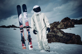 A person in a white spacesuit stands on snowy ground next to two Icelantic Skis Nomad 100 skis featuring a moon design, with rocky terrain and a cloudy sky in the background.