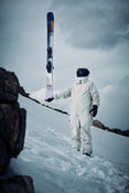 An individual in a white spacesuit and black helmet stands on snowy ground, holding the Icelantic Skis Nomad 112 upright. Rocky terrain is visible in the foreground beneath a cloudy sky.