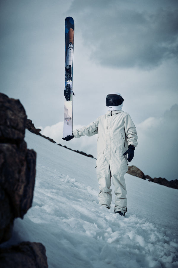 An individual in a white spacesuit and black helmet stands on snowy ground, holding the Icelantic Skis Nomad 112 upright. Rocky terrain is visible in the foreground beneath a cloudy sky.