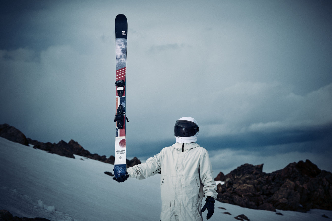 Clad in a white snowsuit and helmet, a person stands on a snowy slope holding an upright Icelantic Skis Nomad 100 ski, with rocky terrain and a cloudy sky in the background.