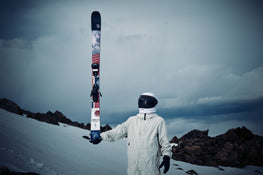 Clad in a white snowsuit and helmet, a person stands on a snowy slope holding an upright Icelantic Skis Nomad 100 ski, with rocky terrain and a cloudy sky in the background.