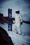 Wearing a white snowsuit and helmet, a person stands on a snowy slope next to the Icelantic Skis Nomad 94, with rocky terrain and a cloudy sky in the background.