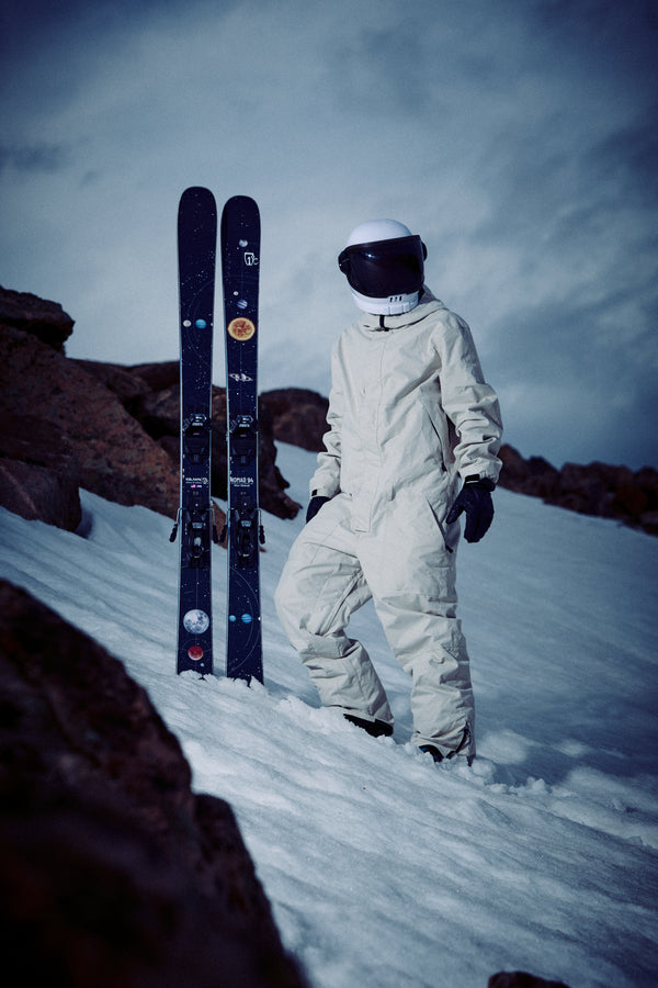 Wearing a white snowsuit and helmet, a person stands on a snowy slope next to the Icelantic Skis Nomad 94, with rocky terrain and a cloudy sky in the background.