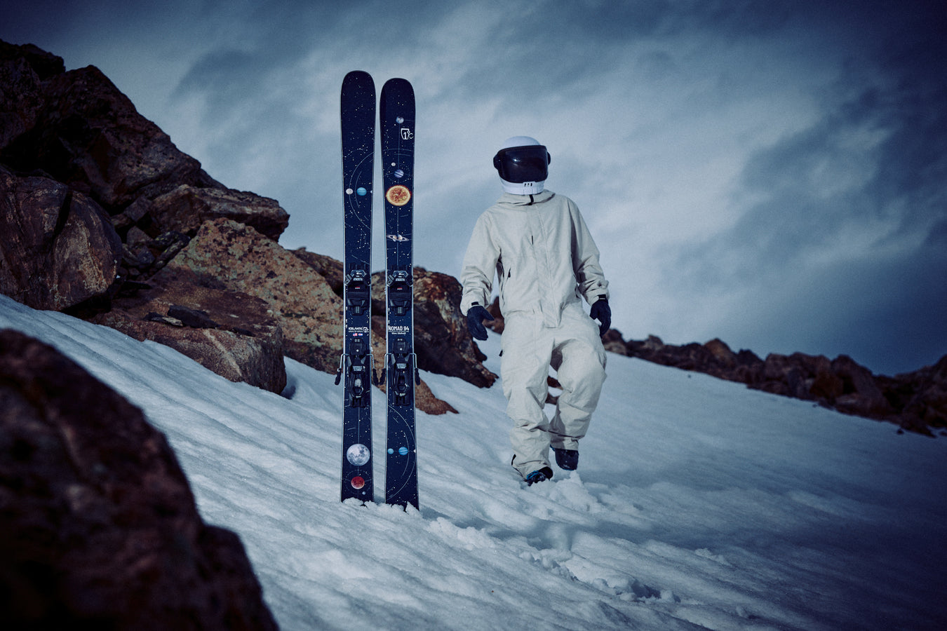A person in a white spacesuit and helmet stands on snowy ground near two upright Icelantic Skis Nomad 94 skis, with rocky terrain in the background under a cloudy sky.