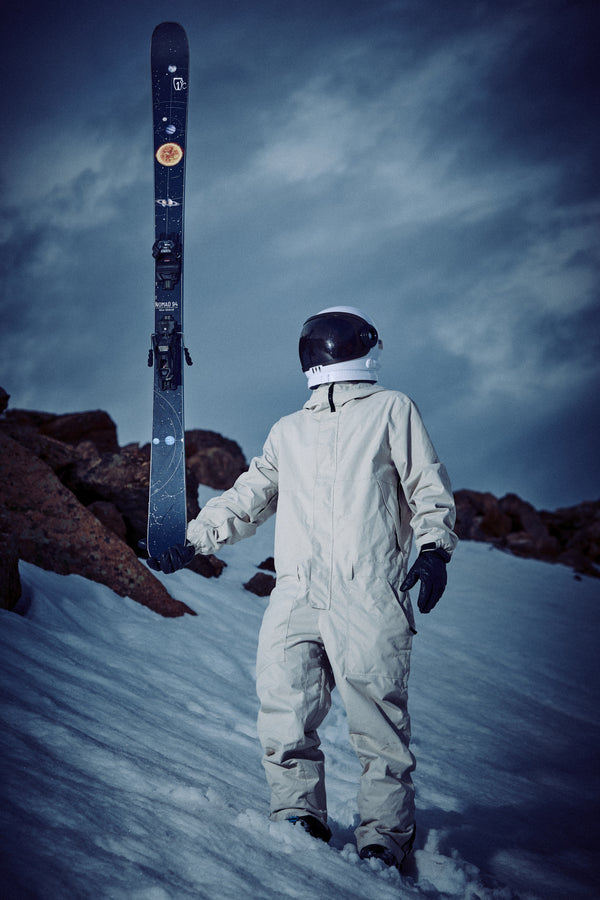 Clad in a white spacesuit, a person stands on snowy ground holding a pair of Icelantic Skis Nomad 94 upright, with rocky terrain and a cloudy sky in the background.
