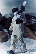 Clad in an astronaut suit, a person carries the Icelantic Skis Nomad 106 on their shoulder while walking across snow-covered, rocky terrain beneath a cloudy sky.